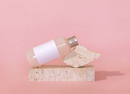 Bottle with blank label lies lies on travertine stone on light pink, close up, mockup. Skincare beauty product. Natural cosmetics, lotion or cleanser. Minimal compositionの写真素材