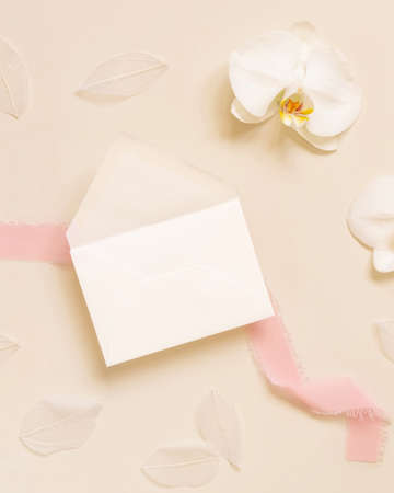 Wedding envelope near white orchid flower and silk ribbons on light yellow top view, mockup. Tropical scene with horizontal blank card flat layの写真素材