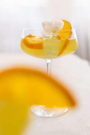 Transparent cocktail with ice cubes in a champagne coupe glass decorated with slices of orange and white orchid flower close up. Tropical drink at the bar. Summer vacation moodの写真素材