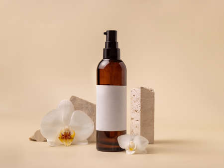 Dark glass cosmetic bottle with blank label near white orchid flowers and stones on light yellow, close up, mockup. Skincare handmade beauty product, tonic or lotion. Exotic natural cosmeticsの写真素材