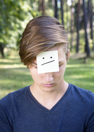 On the forehead shows (drawn) confuse icon on yellow stickers. Concept of negative thoughts and emotions. Confused boy is sitting on green grass in nature.の写真素材