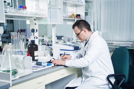 Male lab worker with glasses and white coat with pencil writing down observations in laboratory.の写真素材