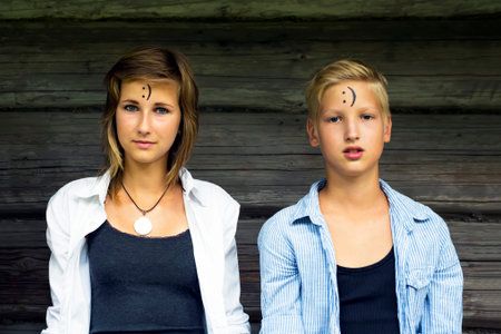 Boy and girl (siblings) are sitting  on a dark wooden background. Neutral look. On the forehead shows (drawn) smile icon. Concept of positive thoughts and emotions.の写真素材