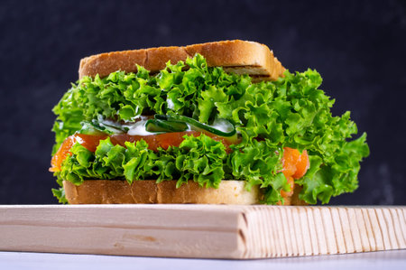 Sandwich with fresh salmon, lettuce, cucumber and sauce on a cutting board, on a dark background....