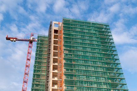 Close-up of a construction site with scaffolding and green safety netting, showcasing an unfinished high-rise building with exposed concrete and brick walls.の写真素材