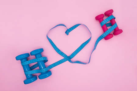 Composition with blue and pink dumbbells and a tailors centimeter in the form of a heart on a pink background. Sports activities for two. Valentines day concept in fitness club.の写真素材