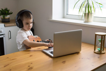 Concentrated boy in headphones plays ukulele guitar and watches online course on laptop while...