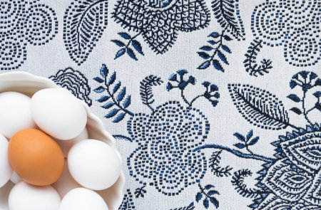 Many white eggs and one brown egg in a white dish against the background of a patterned tablecloth....