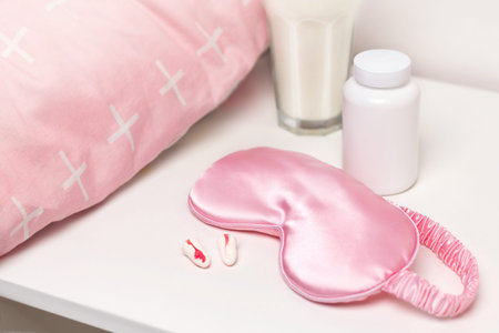 Bedside table with a silk pink sleep mask, earplugs, warm milk for a good night's sleep and sleeping pill mock up. All conditions for good deep sleep. Help for falling asleep free spaceの写真素材
