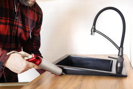 A man installs a black stone sink into a kitchen countertop. Worker seals up the kitchen sink with a sealant using a construction sealing gun close upの写真素材