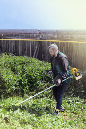 Man mows and cuts the grass with a gasoline trimmer in the garden. Mows nettle grass. vertical photo side viewの写真素材