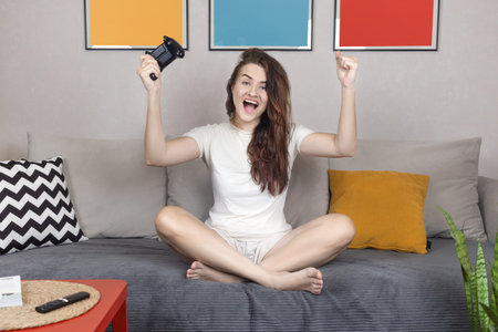 Excited caucasian woman with joystick sitting on couch at home, making victory gesture, winning in...