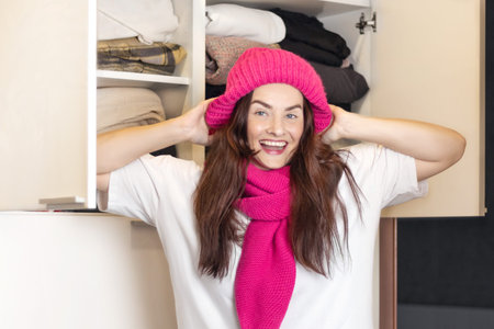 Woman smiles, she found her favorite knitted pink hat and scarf in the closet. Tries on himself, waiting for autumn, winter to wear warm clothes, close up photoの写真素材