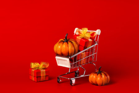 Supermarket trolley, cart with red gifts with gold bows and orange pumpkins. The concept of buying gifts for holidays and sales. Happy Halloween shoppingの写真素材