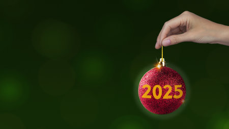 Red Christmas ball with yellow 2025 in woman's hand on green background, horizontal New year style photo banner copy spaceの写真素材