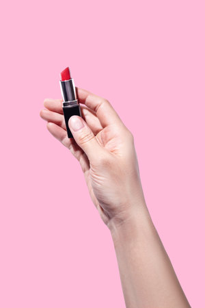 A woman's hand holds red lipstick isolated on pink background vertical photoの写真素材