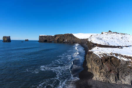 Aerial winter landscape view of Dyrholaey, Iceland. Beautiful winter view of picturesque peninsula with black sand beach, lighthouse and large arch of volcanic rocks in the ocean.の写真素材