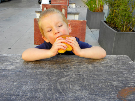 Portrait of a charming blond boy. A three year old boy sits at a wooden table and eats a bun.