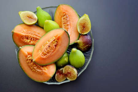 Slices of ripe and juicy cantaloupe melon and sweet figs in a round plate on a gray background. A plate with autumn fruits.の写真素材