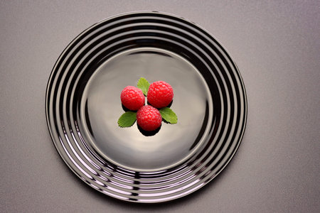 Red raspberries and green mint leaves in a black plate on a gray background.