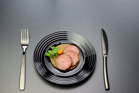 Three slices of salami and green parsley leaves on a slice of round rye bread in a black plate on a gray background.の写真素材