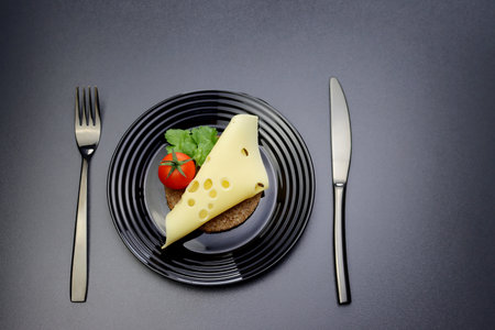 A slice of maasdam cheese, cherry tomatoes and green parsley leaves on a slice of round rye bread in a black plate on a gray background.の写真素材