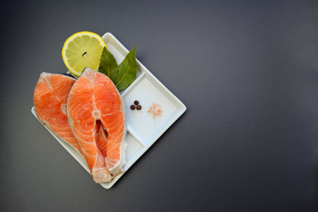 Two fresh salmon steaks, lemon and spices in a square plate on a gray background.の写真素材