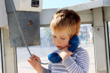A sad boy stands in a telephone booth. A blond boy holds a blue telephone receiver in his hand with his eyes down.の写真素材