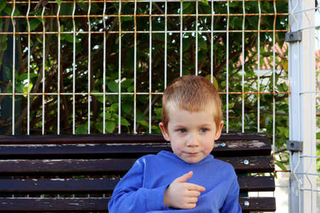 The blond boy looks into the distance thoughtfully. A serious four-year-old boy sits on a bench.の写真素材