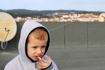A blond boy with a stained chin puts his finger in his mouth. A four-year-old boy in the hood and a dirty faceの写真素材