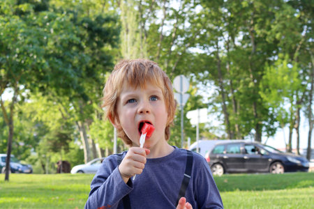 The blond boy holds a fork in his hand and eats a watermelon. A five-year-old boy narrows his eyes...