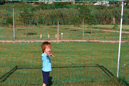 A blond boy is standing near the football goal with his hand outstretched forward. A five-year-old boy stands sideways and squints his eyes.の写真素材