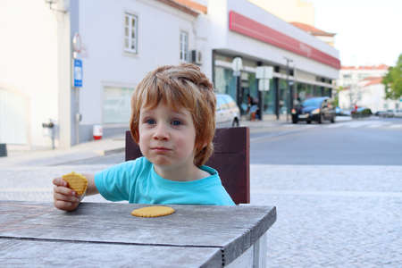 A blond boy sits at a wooden table in a street cafÃ© eating cookies. The five year old boy is eating and smiling.の写真素材
