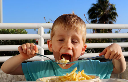 The blond boy with his eyes closed, opens his mouth wide and puts food in it. A five-year-old boy with a fork and knife near his mouth.の写真素材