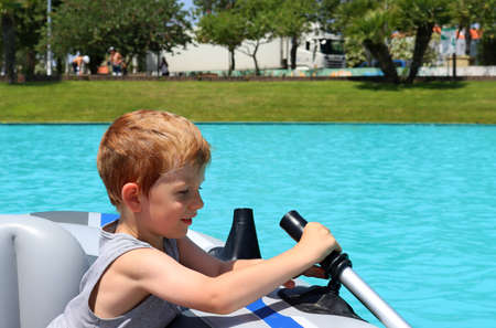 A blond boy sits in a rubber boat and holds an oar. A five-year-old boy is learning how to steer the boat.の写真素材