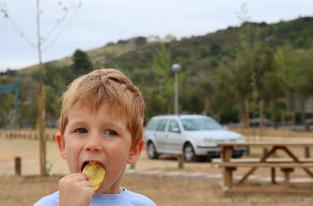 The blond boy opens his mouth and puts chips in it. A five-year-old boy eats junk food.の写真素材