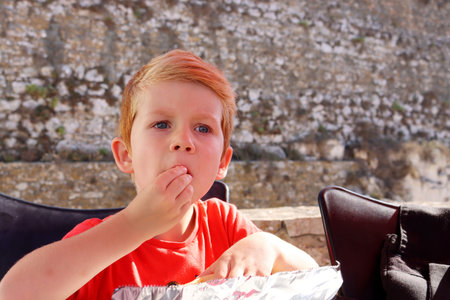 The blond boy puts food in his mouth with his hands. A five-year-old boy eats lunch at a street cafÃ©.の写真素材