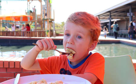 The blond boy eats pasta and looks intentionally into the distance. A five-year-old boy eats lunch at a street cafÃ©.の写真素材