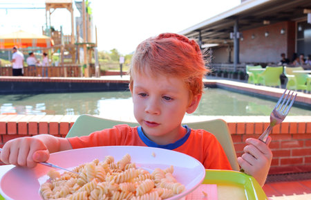 The blond boy looks intentionally at the plate of pasta. A five-year-old boy with a knife and fork sits at the table.の写真素材