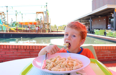 The blond boy opens his mouth and puts macaroni in it. A five-year-old boy eats lunch at a street cafÃ©の写真素材