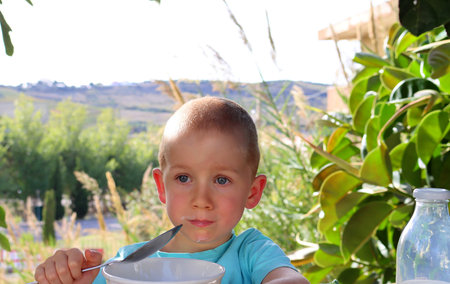 A short-cut, blond boy with a milky mustache stares thoughtfully into the distance. A five-year-old boy eats his lunch outdoors.の写真素材