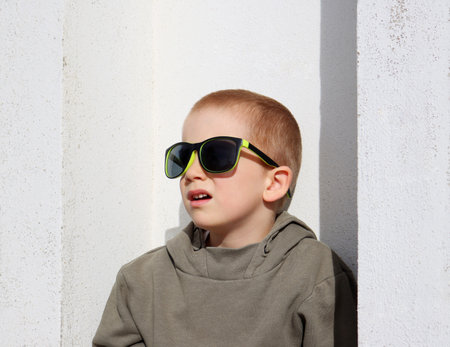 A short-cut, blond boy with sunglasses against a shabby white wall. A five-year-old boy imagines himself as an adultの写真素材