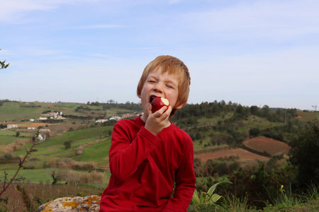 The blond boy screwed up his eyes and bites off a red apple. A five-year-old boy eats fruit outdoors.の写真素材