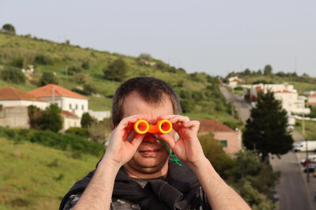 A bristly man looks through children's orange binoculars. Young man with binoculars in the background of the village.の写真素材