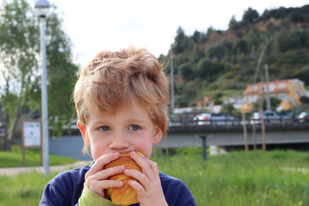 The blond boy takes a bite of a croissant and looks thoughtfully into the distance. Serious five year old boy.の写真素材