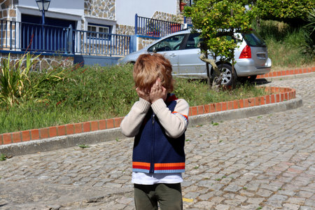 A red-haired boy stands alone in the middle of the street and covers his face with his hands.の写真素材