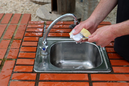 Male hands hold a soap sponge and a spoon over the sink. Washing dishes in the outdoor kitchen.の写真素材