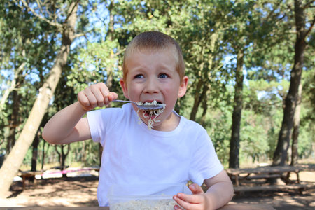 A blond boy brings a spoonful of noodles to his mouth. Six year old boy having lunch in the park on a summer dayの写真素材
