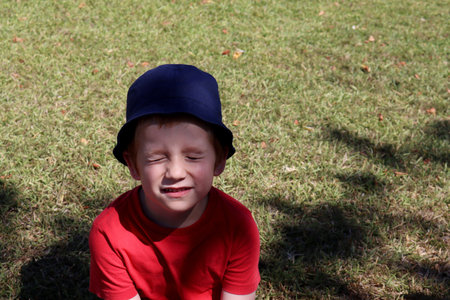 Emotional boy in a red t-shirt and blue cap. A six-year-old boy closed his eyes as he sat on the green grass.の写真素材