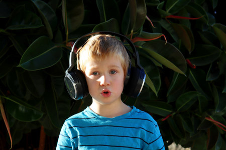 A blond boy listens to music in headphones against the background of green plants. A six year old boy hums a song.の写真素材
